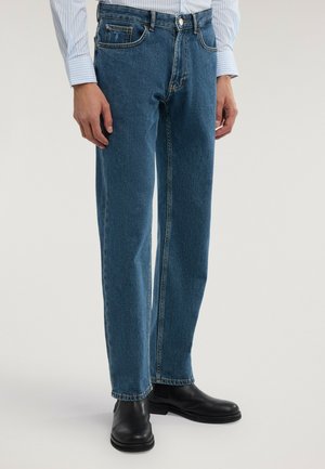 Man som bär raka blå jeans, svarta skinnstövlar och en ljusblå randig långärmad skjorta med händerna avslappnade vid sidorna.