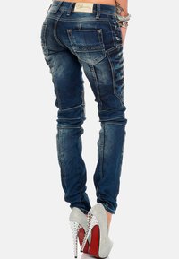 Jeans attillati blu scuro con una finitura rovinata, dotati di tasche posteriori e cuciture a contrasto, abbinati a scarpe con tacco alto argentate.