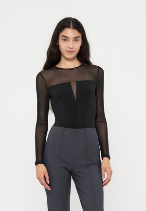 Donna con lunghi capelli scuri in piedi frontalmente, indossa una maglia nera trasparente a maniche lunghe e pantaloni grigi a vita alta su uno sfondo semplice.