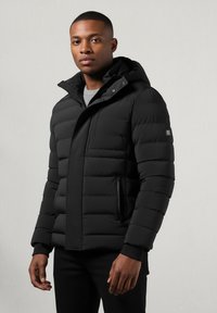 Juodas pūkinis švarkas su užtrauktuku priekyje, gaubtu ir raišteliais ant rankovių. Yra siuvinėjimo detalės ir šoninės kišenės. Medžiaga atrodo lygi ir izoliuota.