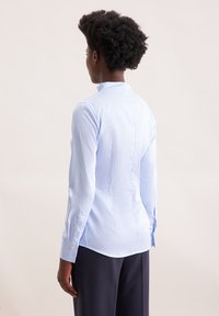 Persona con capelli corti e ricci, che indossa una camicia a righe blu chiaro e pantaloni scuri, vista di spalle su uno sfondo neutro.