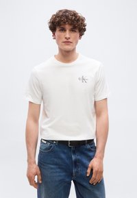 Νεαρός με σγουρά μαλλιά, φορώντας λευκό μπλουζάκι Calvin Klein και μπλε τζιν, στέκεται μπροστά σε απλό φωτεινό φόντο.