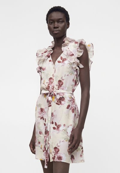 Γυναίκα που φορά ένα αμάνικο, μέχρι το γόνατο φόρεμα με λουλούδια, βολάν στους ώμους και ζώνη στη μέση, στέκεται μπροστά σε ένα μονόχρωμο φόντο.