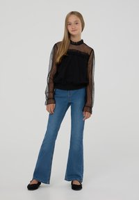 Blusa trasparente nera a pois con collare e polsini arricciati, abbinata a jeans in denim blu a zampa d'elefante e ballerine nere. Outfit semplice e casual.