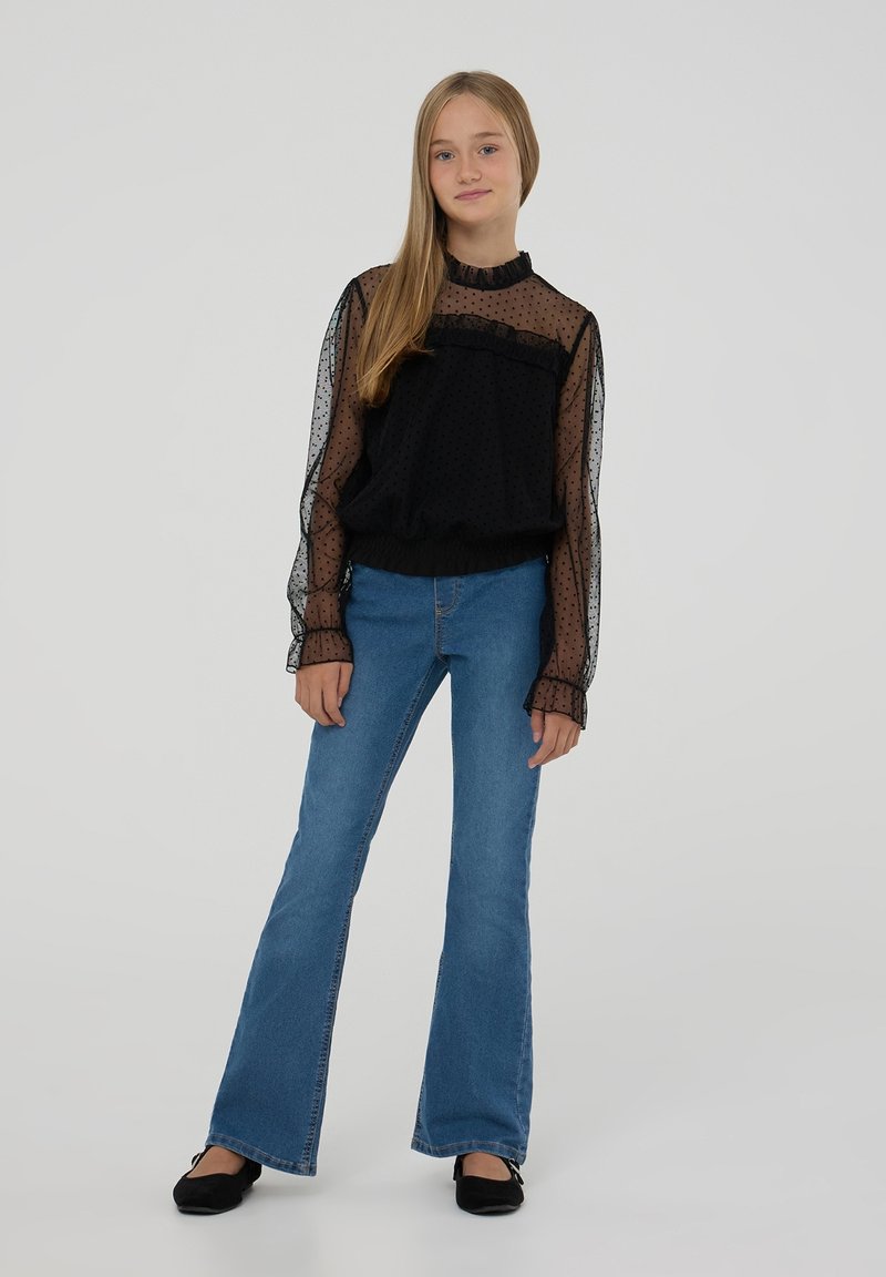 Blusa trasparente nera a pois con collare e polsini arricciati, abbinata a jeans in denim blu a zampa d'elefante e ballerine nere. Outfit semplice e casual.