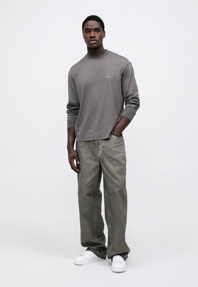 Pelēks garām piedurknēm krekls ar mazu logotipu, apvienots ar brīvām olīvu zaļām biksēm un baltām sporta kedām, uz vienkārša fona.