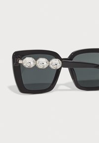 Óculos de sol retangulares pretos com lentes escuras, apresentando três grandes strass claros na parte superior e uma armação brilhante.
