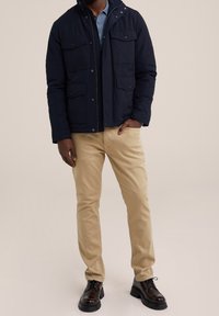 Man som bär en marinblå jacka, blå skjorta, beige byxor och mörkbruna läderskor med snörning, står med en hand i fickan.