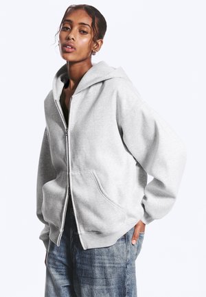 Jonge vrouw draagt een lichtgrijze hoodie met rits en blauwe spijkerbroek, staat met één hand in haar zak tegen een effen achtergrond.