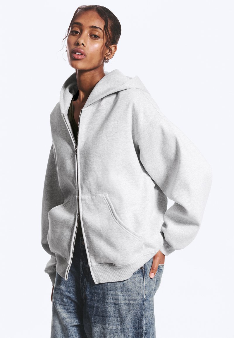 Giovane donna che indossa una felpa con cappuccio grigio chiaro con zip e jeans blu, in piedi con una mano in tasca su uno sfondo semplice.
