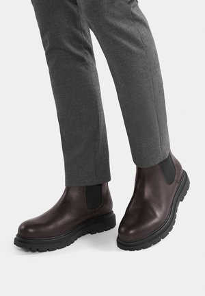 Smeđe Chelsea čizme od kože s elastičnim bočnim panelima, crnim gumenim potplatom i teksturiranim završetkom, uparene sa sivim kariranim hlačama.