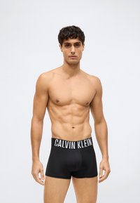 T-paitta paitsi mies, jolla on lyhyet kiharaiset hiukset, mallintaa mustia Calvin Klein -bokserihousuja vaalean harmaata taustaa vasten.