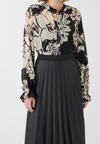 Blouse semi-transparente à motif floral beige et noir, avec un col froncé et une fermeture à boutons, assortie à une jupe plissée noire.