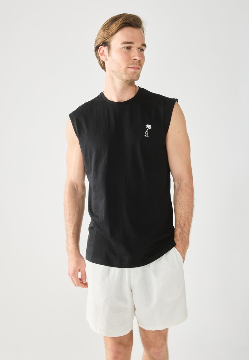 T-shirt sans manches noir avec un petit graphique de palmier blanc sur la poitrine gauche, associé à un short en lin de couleur claire.
