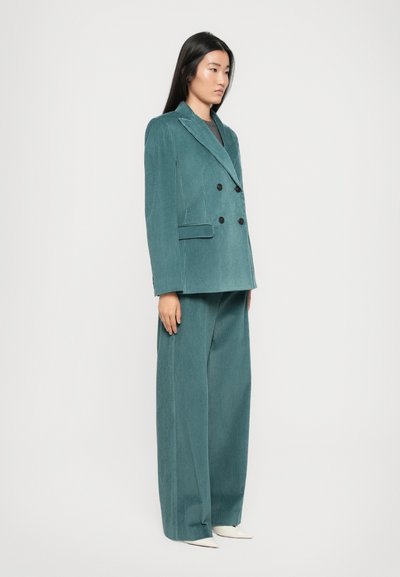 Donna in piedi che indossa un blazer doppiopetto in corduroy teal e pantaloni larghi abbinati, con scarpe a punta bianche su sfondo bianco.