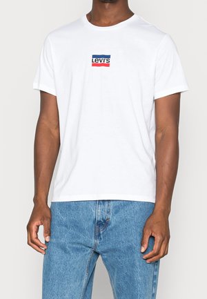 Mand iført hvid Levi's t-shirt med blå og rød logo, kombineret med mellemblå denimjeans, stående mod en ensfarvet baggrund.