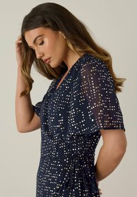 Abito navy con design a wrap, caratterizzato da maniche corte, vita legata e un motivo a Pois metallici sparsi. Tessuto leggero e trasparente.