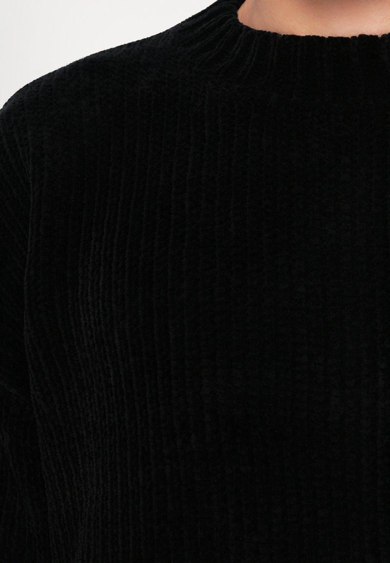Sort fløjlstrik med en tekstureret, ribbet design og høj halsudskæring. Stoffet ser blødt og luksuriøst ud.
