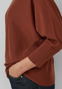Sweter w kolorze rdzy o luźnym kroju, z rękawami do trzech czwartej długości, prążkowanym wykończeniem i gładką fakturą, w połączeniu z ciemnymi dżinsami.