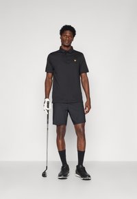 Melnā polo krekls ar īsām piedurknēm, dzeltenais logo uz krūtīm, saskaņots ar melniem šortiem. Golfs tur nūju un valkā melnas kurpes un zeķes.