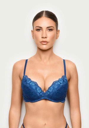 Mujer con cabello castaño con un sujetador azul de encaje mirando hacia adelante sobre un fondo blanco liso, mostrando la parte superior del cuerpo y expresión neutral.
