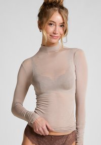 Lichtbeige, lange mouw turtleneck-top in transparante, rekbare stof met geplooide zijkanten, gedragen over een bruine kanten beha en bijpassende kanten onderbroek.