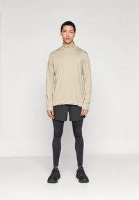 Αθλητική μπλούζα με μακρύ μανίκι, ανοιχτό μπεζ χρώμα, με φερμουάρ, συνδυασμένη με μαύρο σορτς και κολάν. Μαύρα αθλητικά παπούτσια ολοκληρώνουν το σύνολο.