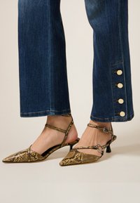 Pies con tacones de punta en estampado de serpiente y correas en el tobillo, combinados con jeans azules oscuros que tienen cuatro grandes botones dorados en el lado.