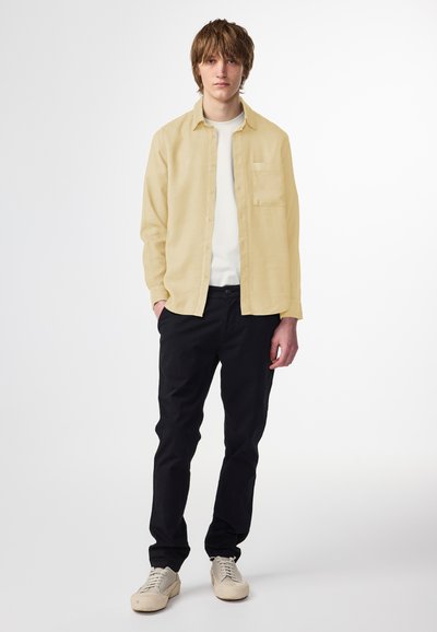 Hellgelbes Hemd über einem weißen T-Shirt, kombiniert mit schwarzen Hosen und beigen Turnschuhen. Einfaches Design mit einer Fronttasche.