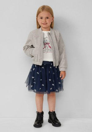 Junges Mädchen mit blonden Haaren, das eine graue Zip-Jacke, ein weißes Shirt mit Katzenmotiv, einen marineblauen Tüllrock mit Schleifen und schwarze Schnürstiefel trägt.