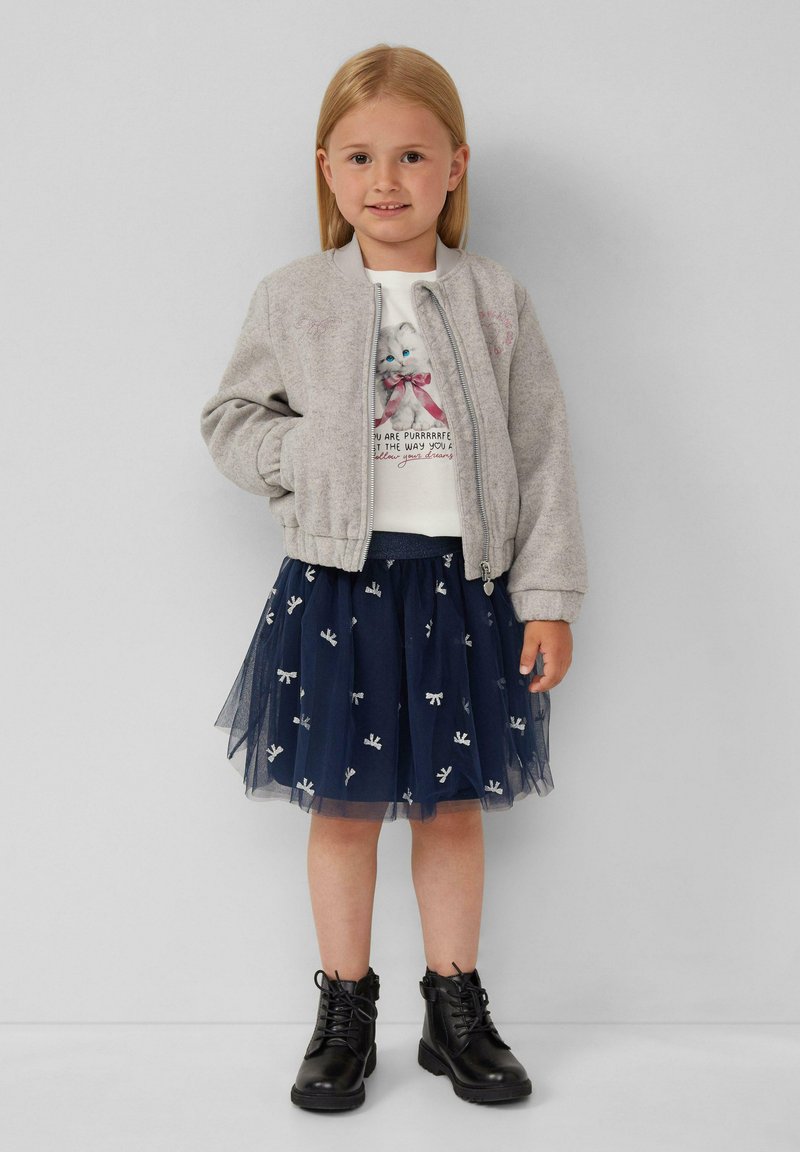 Ragazzina con capelli biondi che indossa una giacca grigia con zip, una maglietta bianca con una grafica di gatto, una gonna di tulle blu navy con fiocchi e stivali neri con lacci.