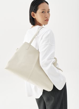 Ženska s kratkimi črnimi lasmi, oblečena v belo majico z dolgimi rokavi in temne hlače, ki nosi veliko bež usnjeno torbo čez ramo.