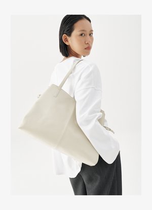 Kvinne med kort svart hår iført hvit langermet topp og mørke bukser, bærende på en stor beige skinnveske på skulderen.