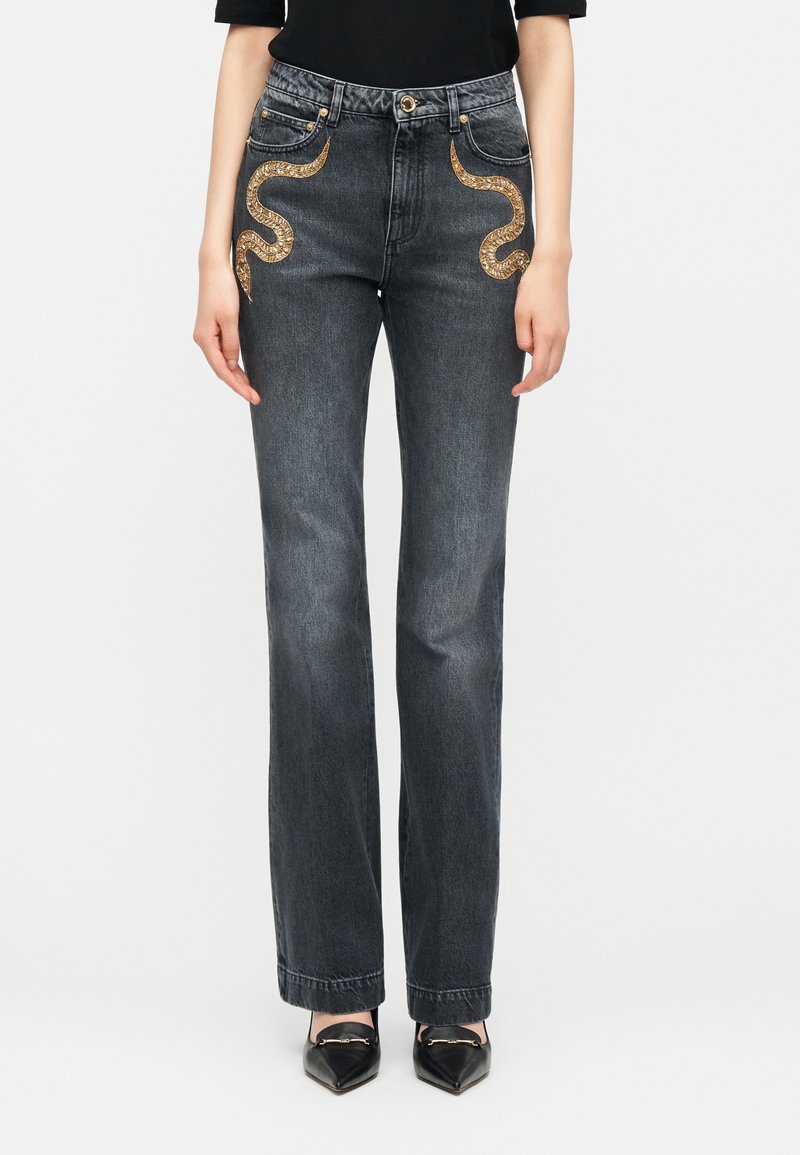 Donkergrijze jeans met rechte pijpen en goudkleurige slangborduursels op de voorzakken, gedragen met zwarte puntige schoenen en een zwart shirt.