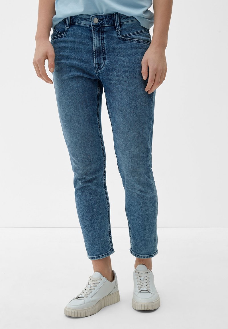 Personne portant un jean bleu taille mi-haute et coupe ajustée avec des baskets blanches à plateforme, debout devant un fond clair uni.