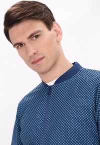 Giacca bomber navy con un motivo a pois in azzurro chiaro, caratterizzata da un colletto alto e una cerniera frontale. Tessuto liscio e design moderno.