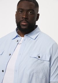 Lichtblauw overhemd met korte mouwen van een gestructureerde stof, met twee poitrinezakken met zwarte knoopaccenten en een contrasterend wit t-shirt eronder.