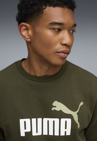 Olivengrøn sweat-shirt med et stort hvidt "PUMA" logo og et lys grøn springende kattegrafik. Glat tekstur og rund hals design.