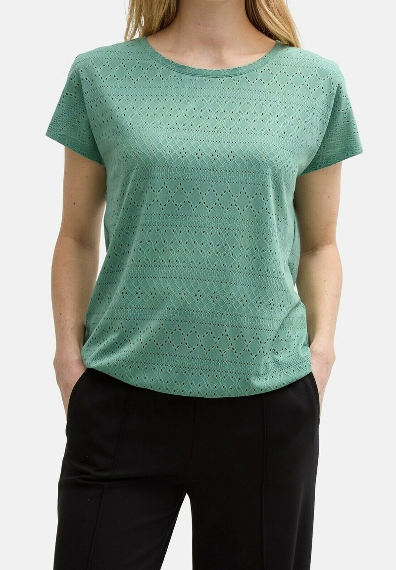 Femme portant un haut vert à manches courtes avec un motif œillet et un pantalon noir, les mains dans les poches, sur un fond uni.