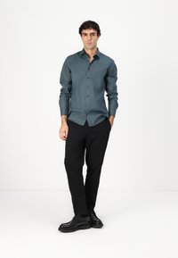 Chemise bleu canard à boutons avec une texture lisse, manches longues, assortie à un pantalon noir et des chaussures noires. Design minimal, silhouette slim.