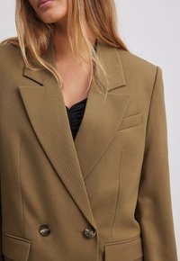 Dwurzędowa oliwkowa marynarka z wyraźnie zarysowanymi ramionami, klapami, dwoma guzikami, teksturowanym materiałem i bocznymi kieszeniami.