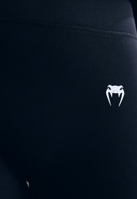 Sorte treningsleggings laget av elastisk stoff, med en hvit edderkopp-logo på siden og ribbet detaljering på midjen.