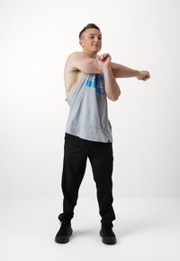 Top cinza sem mangas com gráfico azul, corte solto; calças de treino pretas; sapatilhas pretas; sujeito a esticar o braço com postura relaxada.