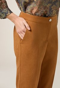 Brązowe spodnie o wysokim stanie, wykonane z gładkiego materiału, z boczną kieszenią, metalowym guzikiem i dopasowanym krojem.