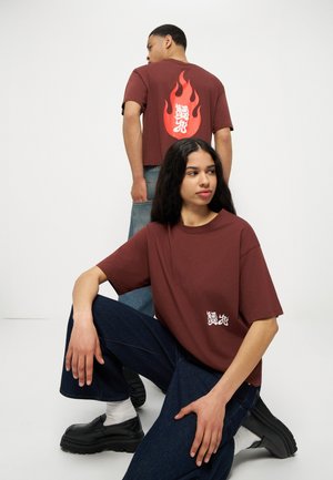 Δύο άτομα που φορούν καφέ T-shirts με λευκό και κόκκινο γραφικό φλόγας κείμενο· το ένα κάθεται στο πάτωμα, το άλλο στέκεται γυρισμένο προς τα πίσω.