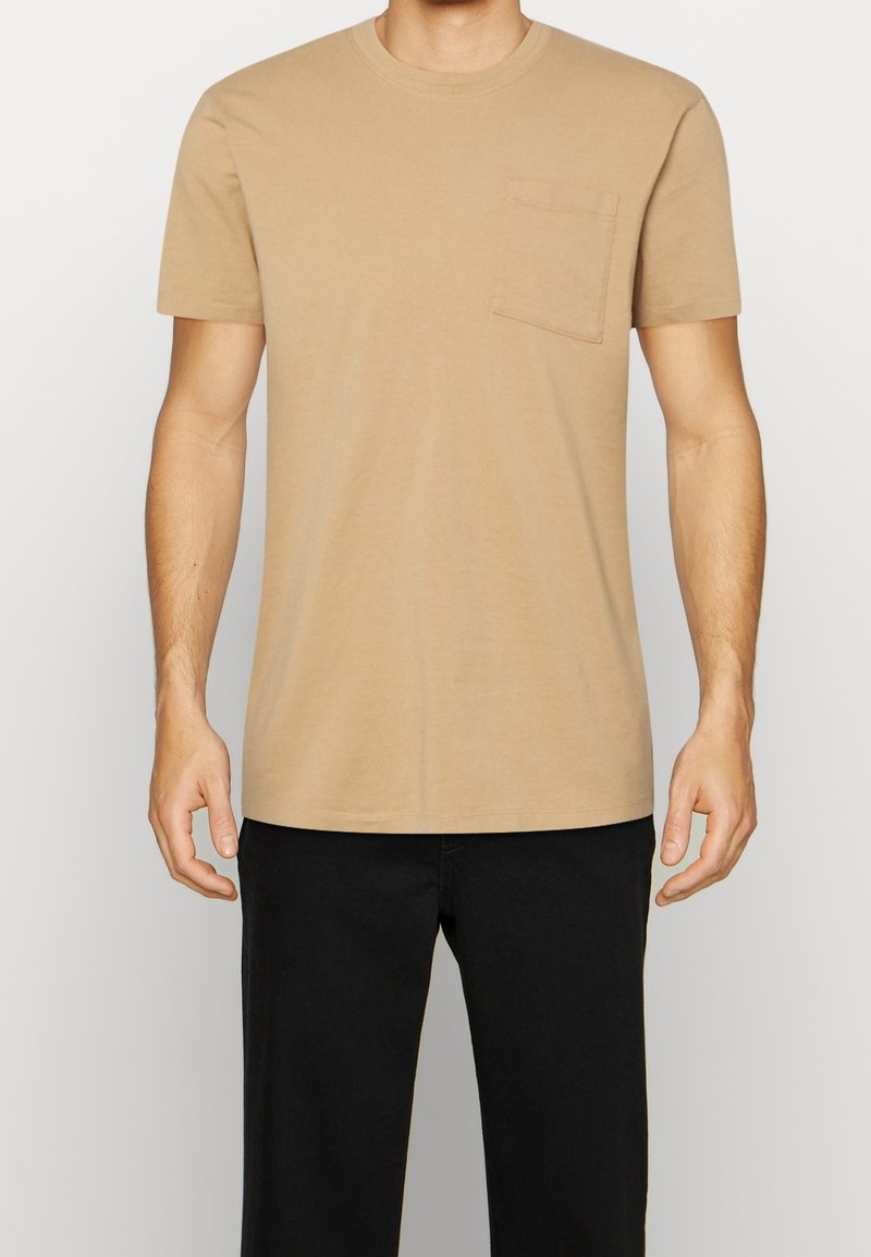 Krémové tričko s krátkým rukávem a kulatým výstřihem, opatřené jednou náprsní kapsou, vyrobené z hladké tkaniny, vystavené na světlém pozadí.