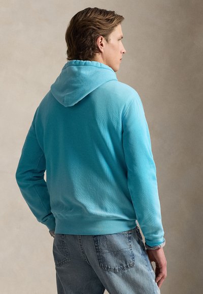 Sweat à capuche turquoise avec une capuche à cordon, ourlet et poignets côtelés, associé à un jean bleu clair. Le tissu semble doux avec un design simple.