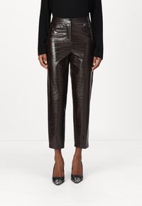 Braune, aus Leder gefertigte, taillierte Hose mit Krokodil-Textur, vorn mit Taschen und einem eleganten, geraden Schnitt. Schwarze High Heels runden den Look ab.