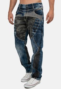 Jean en denim avec un motif géométrique bleu et noir, des accents camouflés, de grands boutons latéraux et des textures contrastées. Portés avec des baskets blanches.