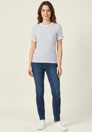 Femme portant une chemise rayée blanche et bleu clair à manches courtes, un jean skinny bleu foncé et des baskets blanches, debout devant un fond uni.